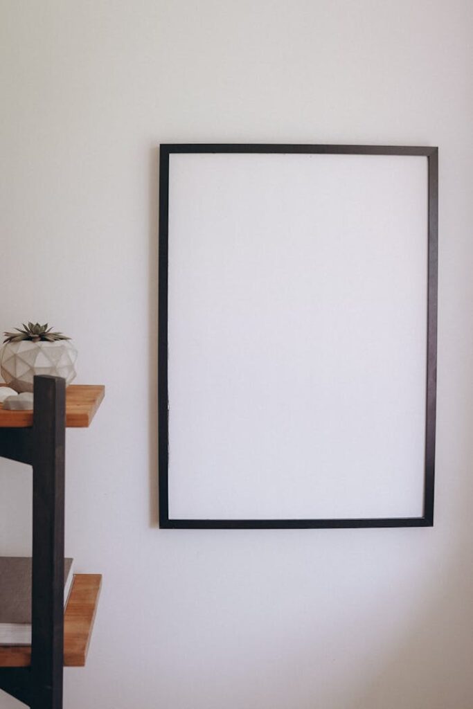 A clean, minimalist blank frame on a neutral-colored wall with a decorative shelf nearby.